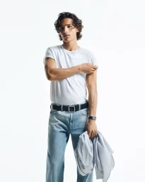Male model wearing a white t-shirt and blue jeans holding a striped shirt against a plain white background