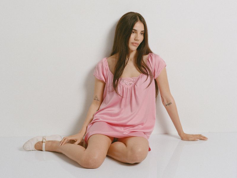 A woman wearing a pink dress and white shoes sitting on a white floor against a plain white wall.