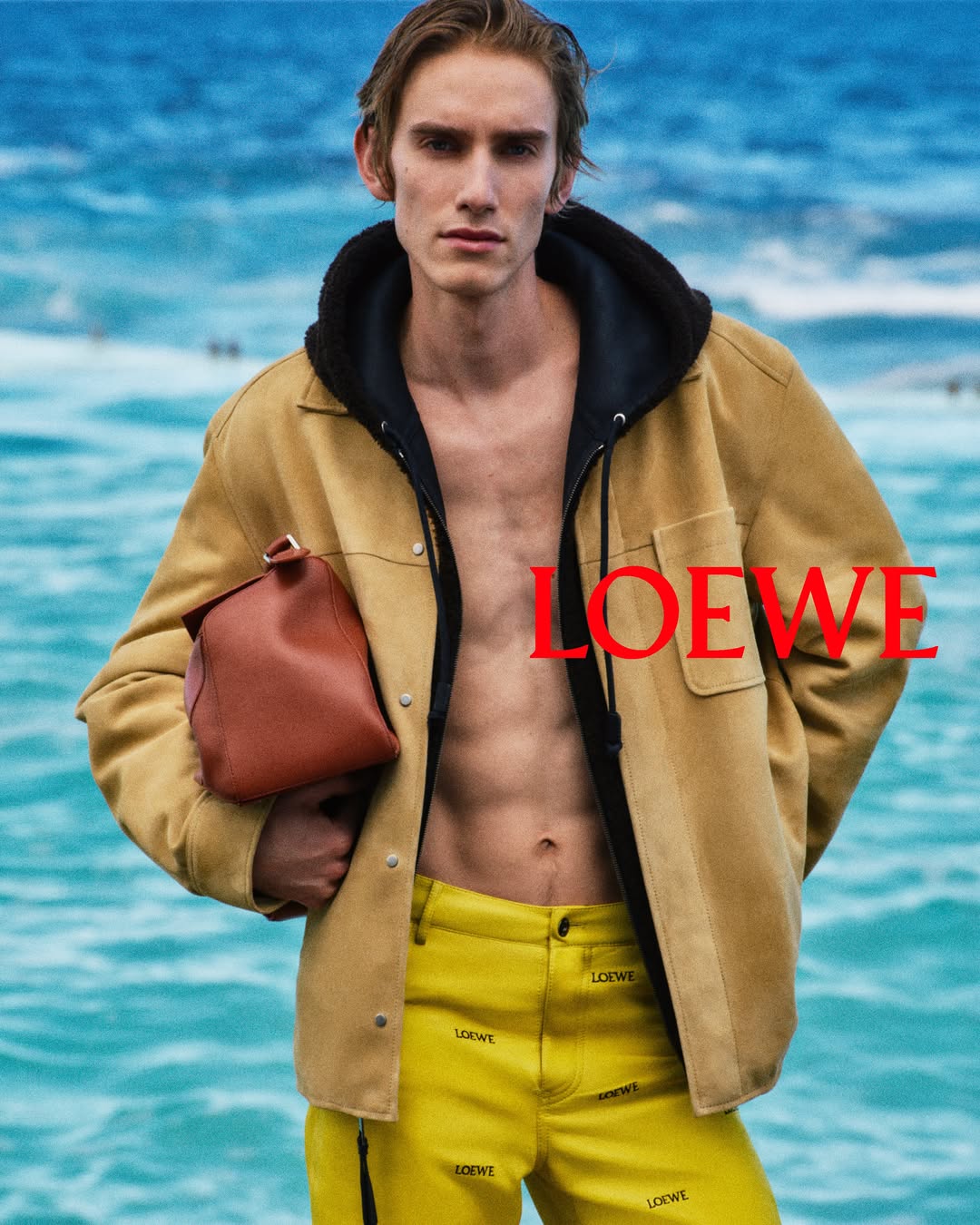 Levon Hawke wearing an open tan suede jacket, yellow logo pants, and holding a brown leather pouch against turquoise ocean waves.