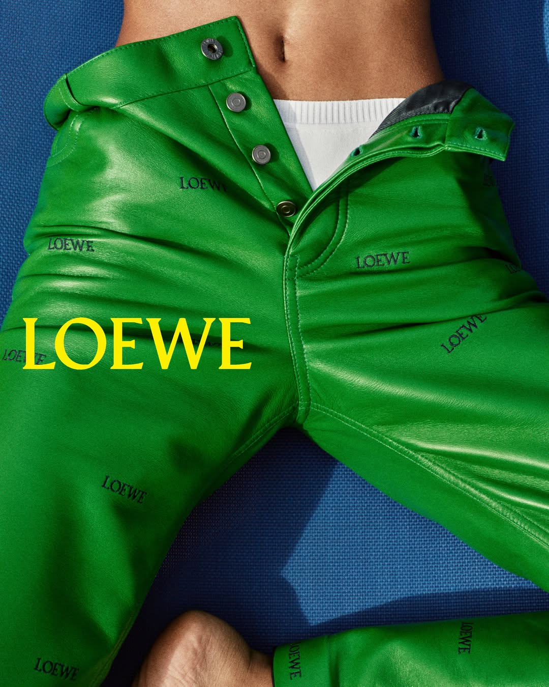 Close-up of vibrant green leather trousers with an unbuttoned fly and subtle embossed logo lettering against a blue surface.