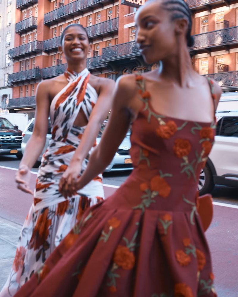 Two women in floral print dresses walk hand-in-hand on a city sidewalk near a red brick building and parked cars, smiling.