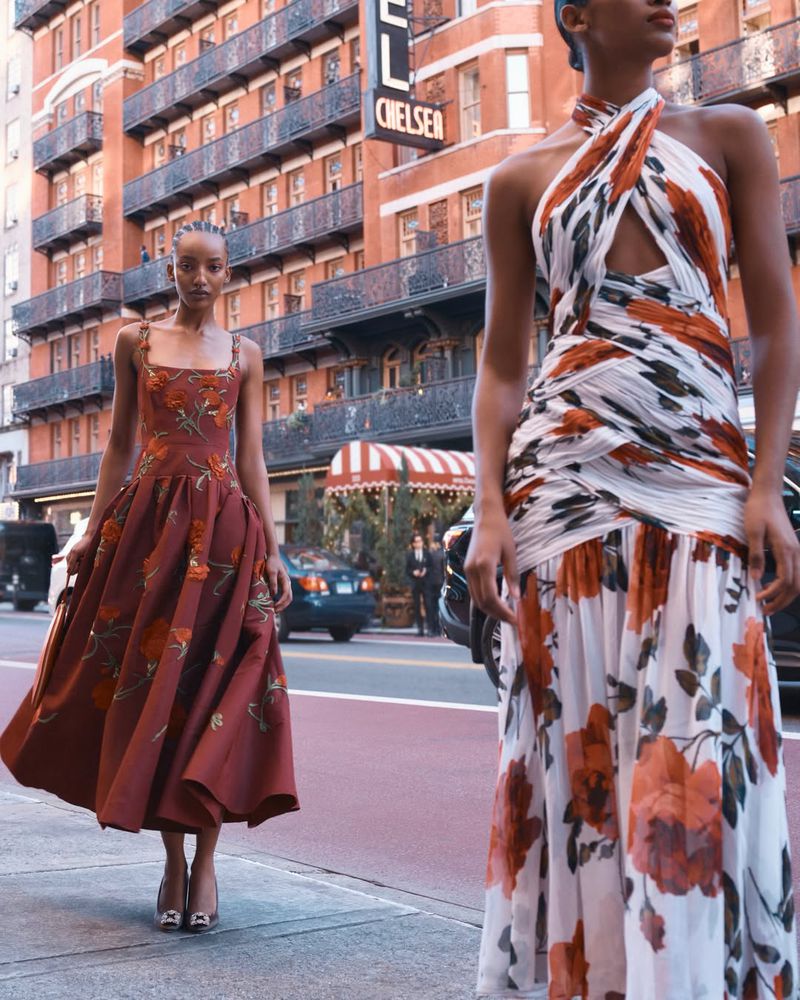 Two female models in floral patterned designer dresses walking along a city street in front of a brick hotel building.