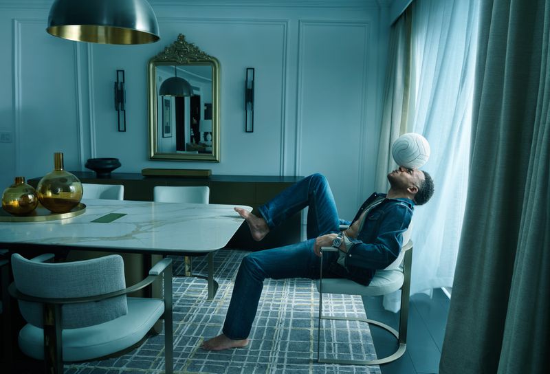 Kylian Mbappe balancing a white football on his forehead while seated in a modern apartment dining room