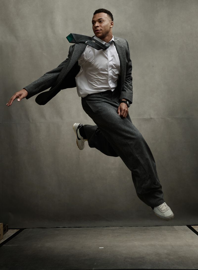 Adult male in a white shirt and grey suit is dynamically posed mid-air in a studio against a grey backdrop, wearing white sneakers.