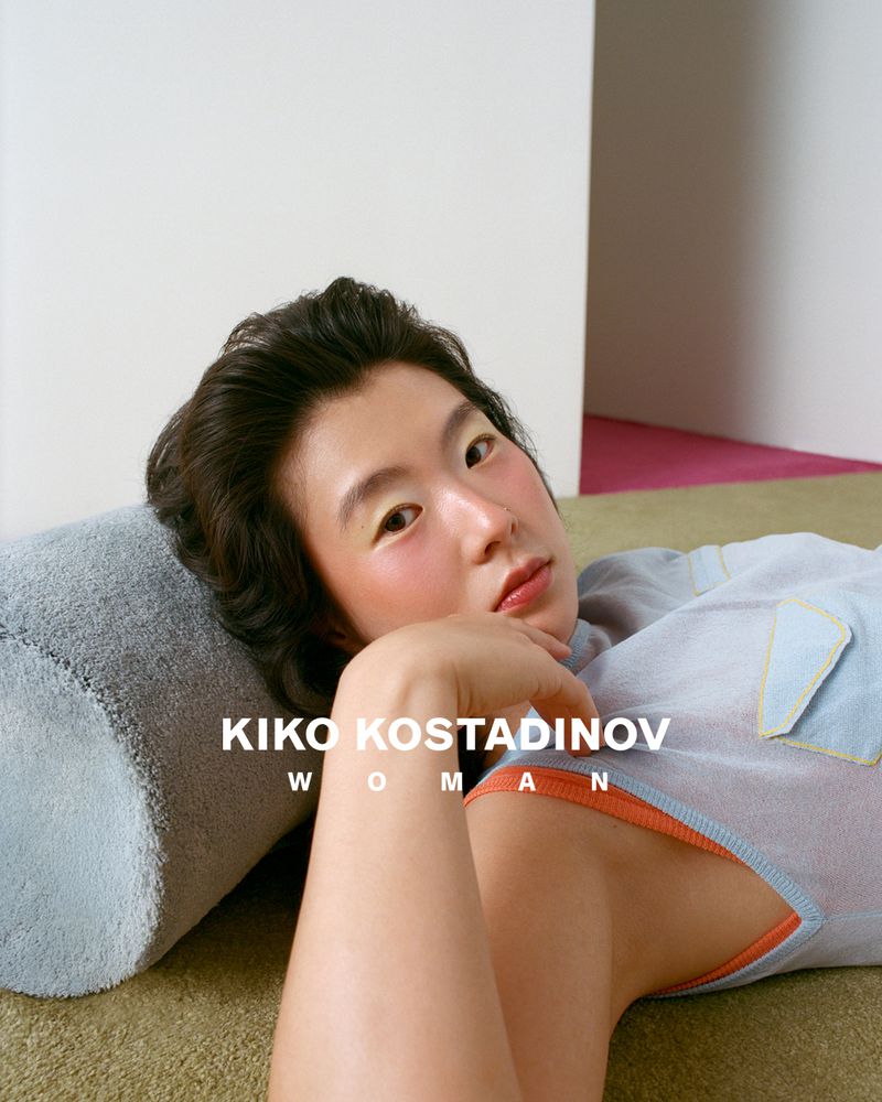 Portrait of an East Asian adult woman with fair skin, lying on a green rug, wearing a light grey top, next to a light blue bolster pillow.