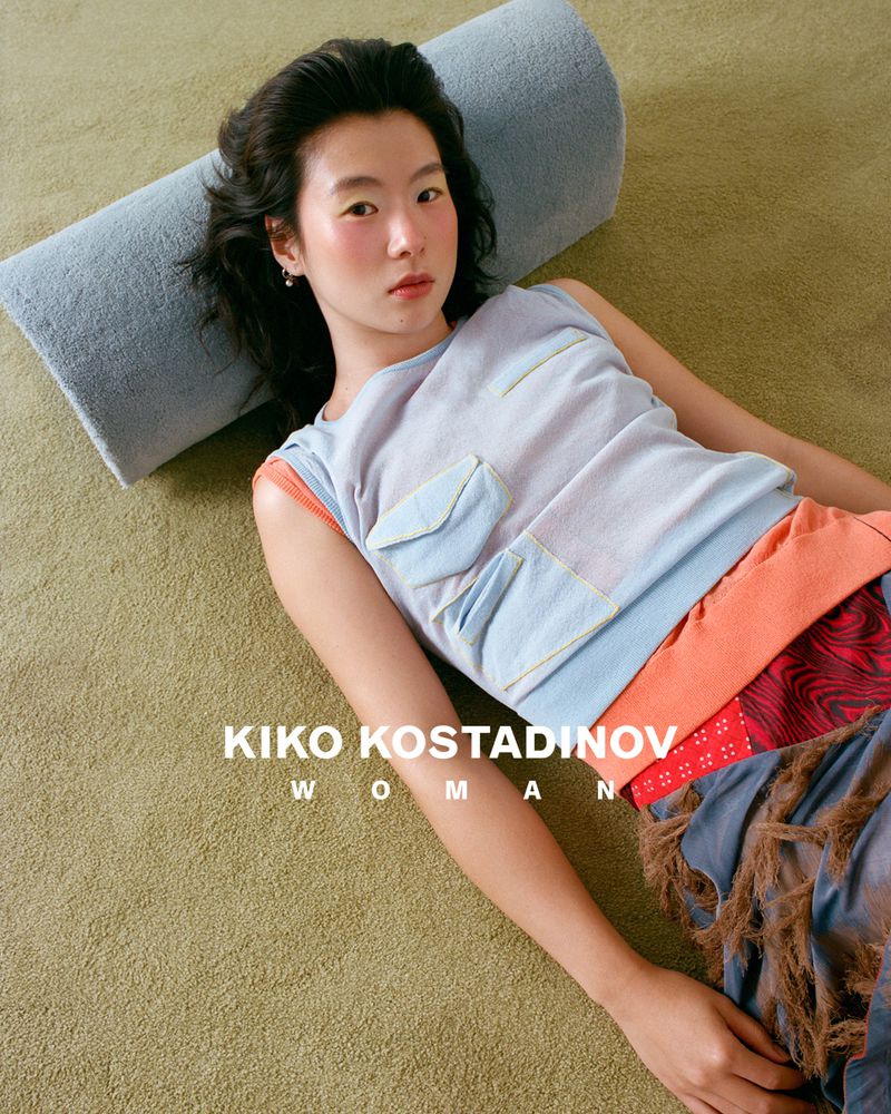 Model lying on a green carpeted floor resting against a grey cylindrical bolster pillow, wearing a blue sleeveless top and patterned skirt.