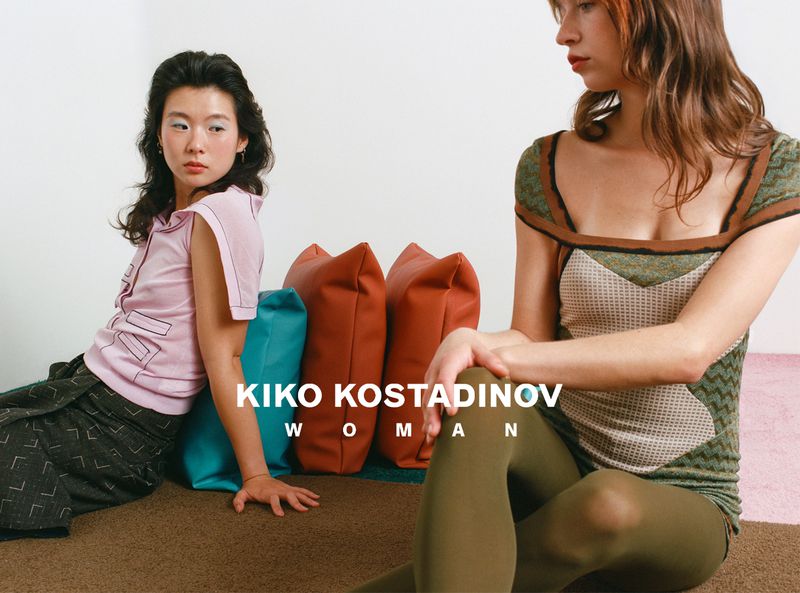 Two female models posing in a minimalist interior with colorful pillows and textured brown carpeting for Kiko Kostadinov SS26.