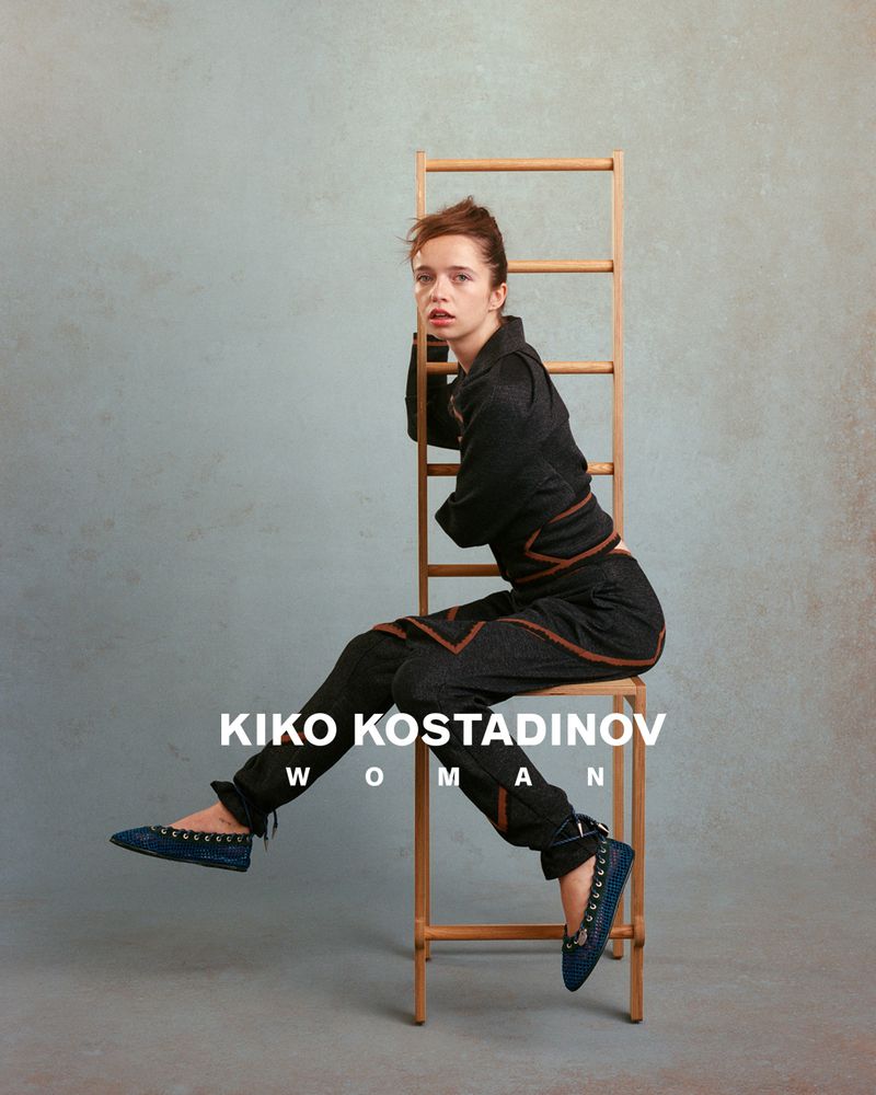 Model seated on a tall wooden ladder chair wearing dark tailored clothing against a textured grey background