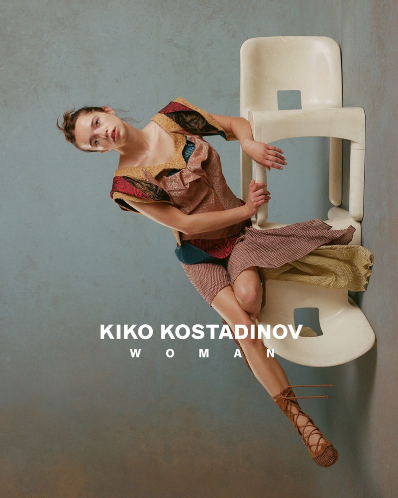 Model wearing patterned designer dress and lace-up shoes posing next to white chairs against textured studio wall for Kiko Kostadinov