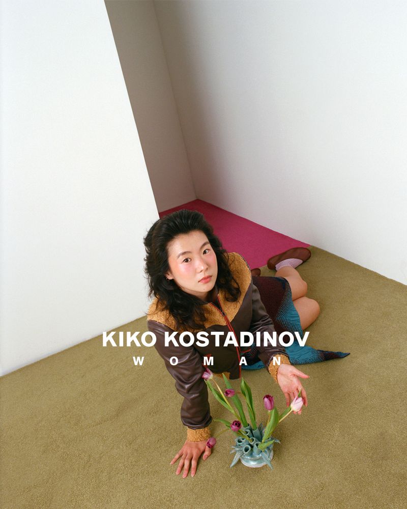 Model in brown jacket lying on a textured olive carpet beside a vase of tulips in a minimalist interior for Kiko Kostadinov SS26.