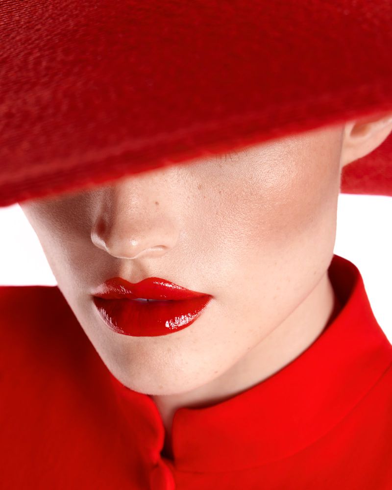 Close-up of a model wearing a red wide-brim hat obscuring the eyes, featuring glossy red lipstick and a matching red collar.