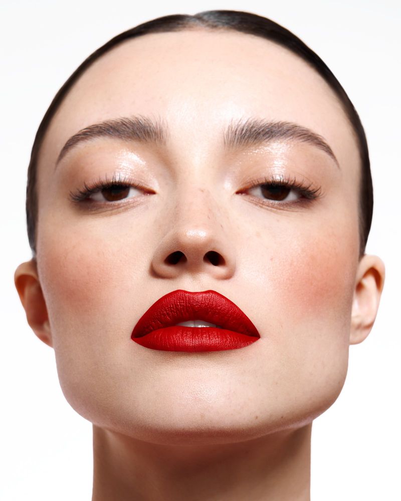 Close-up beauty portrait of a woman with vibrant red lipstick and slicked-back dark hair against a seamless white background.