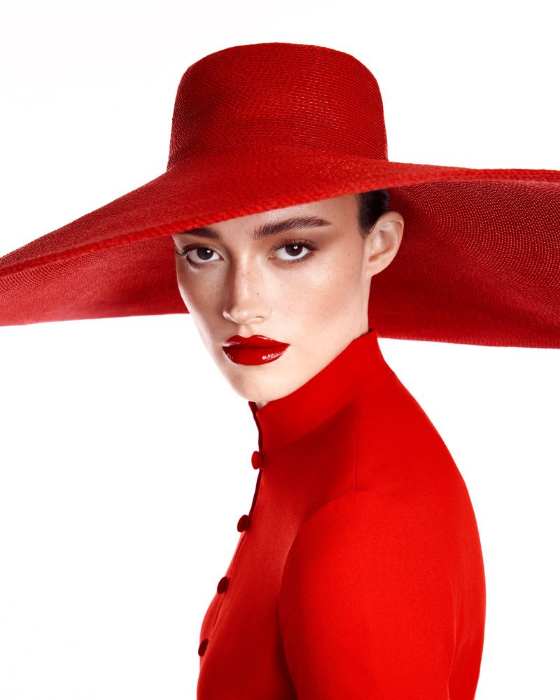 Model wearing a vibrant red wide-brim hat and matching red collared top with glossy red lipstick against a plain white background