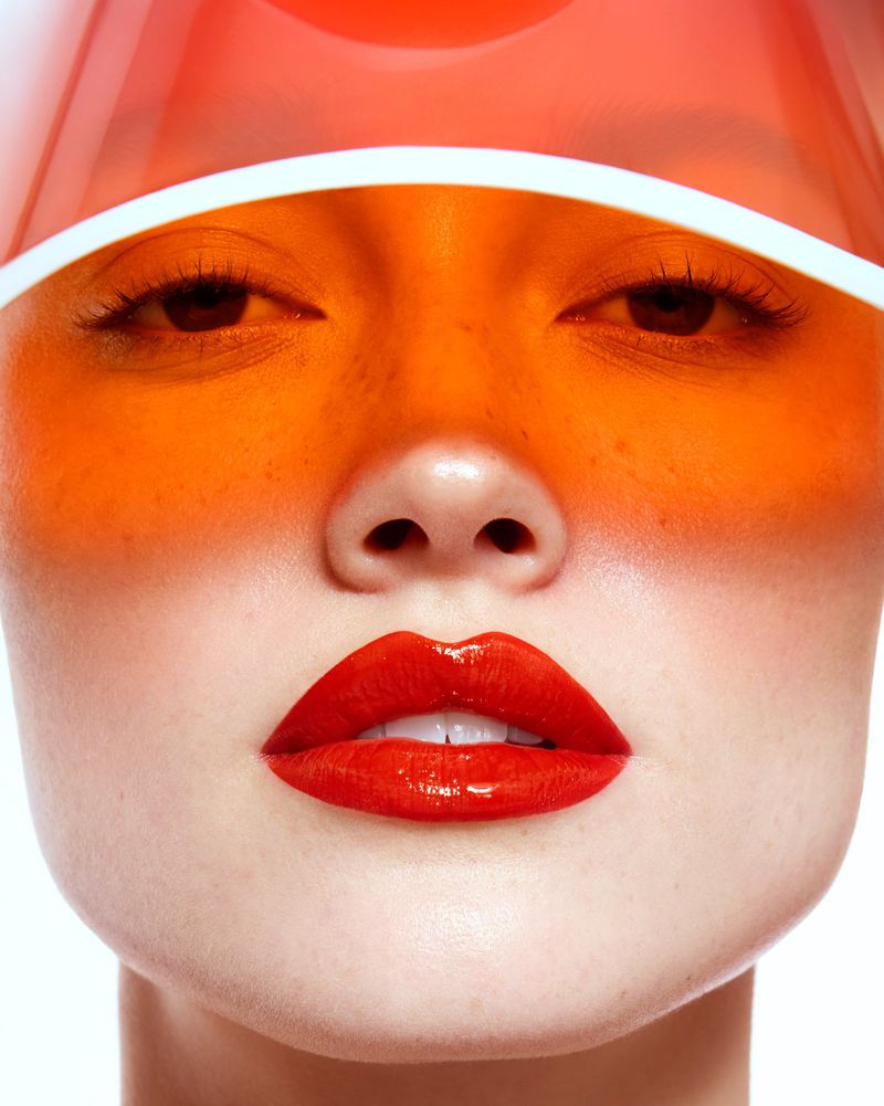 Close-up portrait of a model wearing a translucent orange visor and glossy red lipstick against a plain white background.