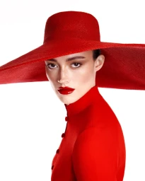 Model wearing a vibrant red wide-brim hat and matching red collared top with glossy red lipstick against a plain white background