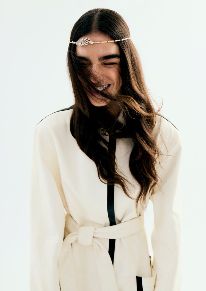 Smiling woman wearing a belted off-white jacket with dark trim and a delicate jewelled headband
