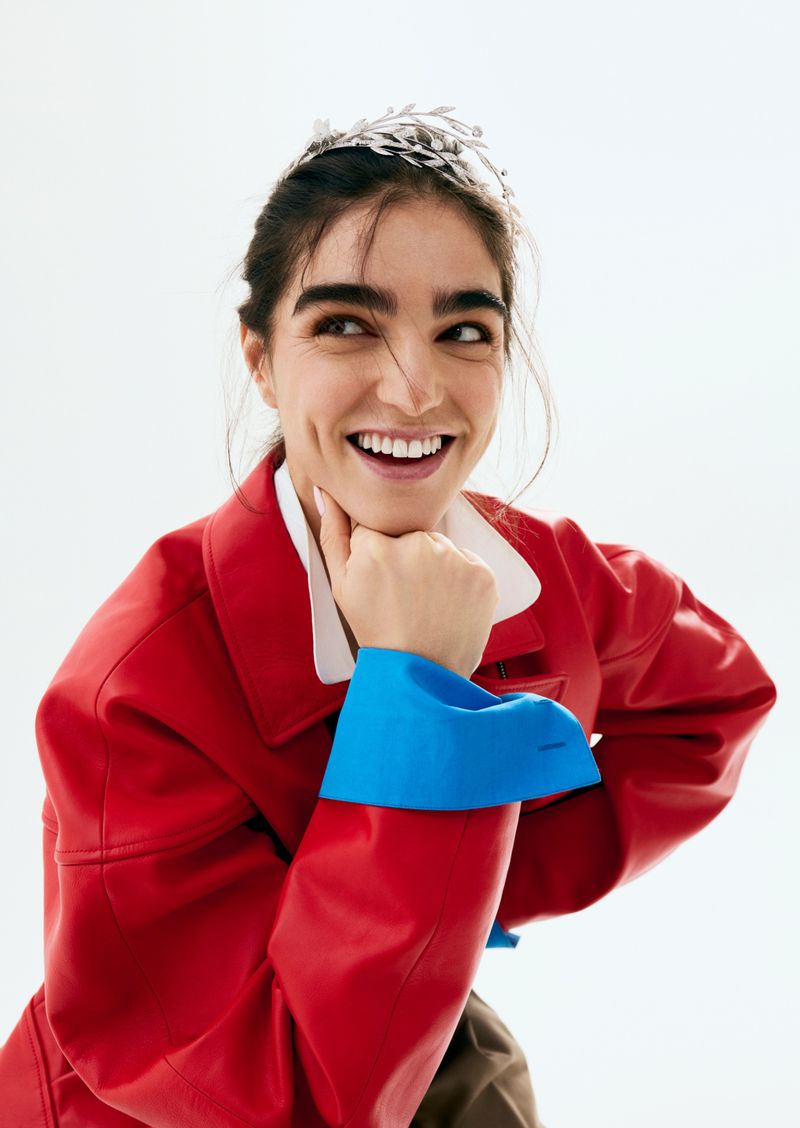 Smiling woman wearing a red jacket with blue cuffs and a metallic headpiece against a white background