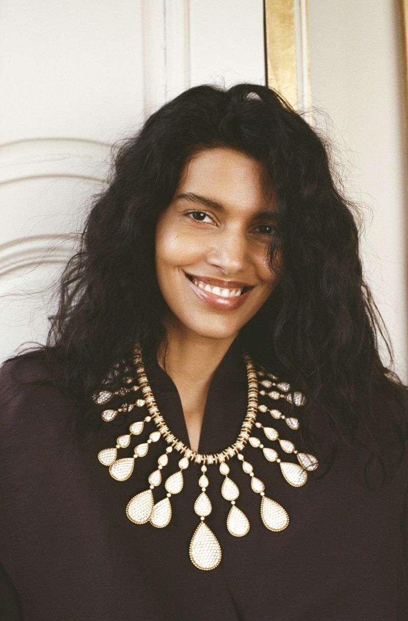 Model Pooja Mor wearing a large statement necklace and dark apparel against a panelled interior background