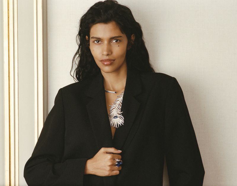 Woman in black blazer poses with luxury jewelry. She wears a large blue ring and an ornate feather necklace. Indoor setting, neutral background.