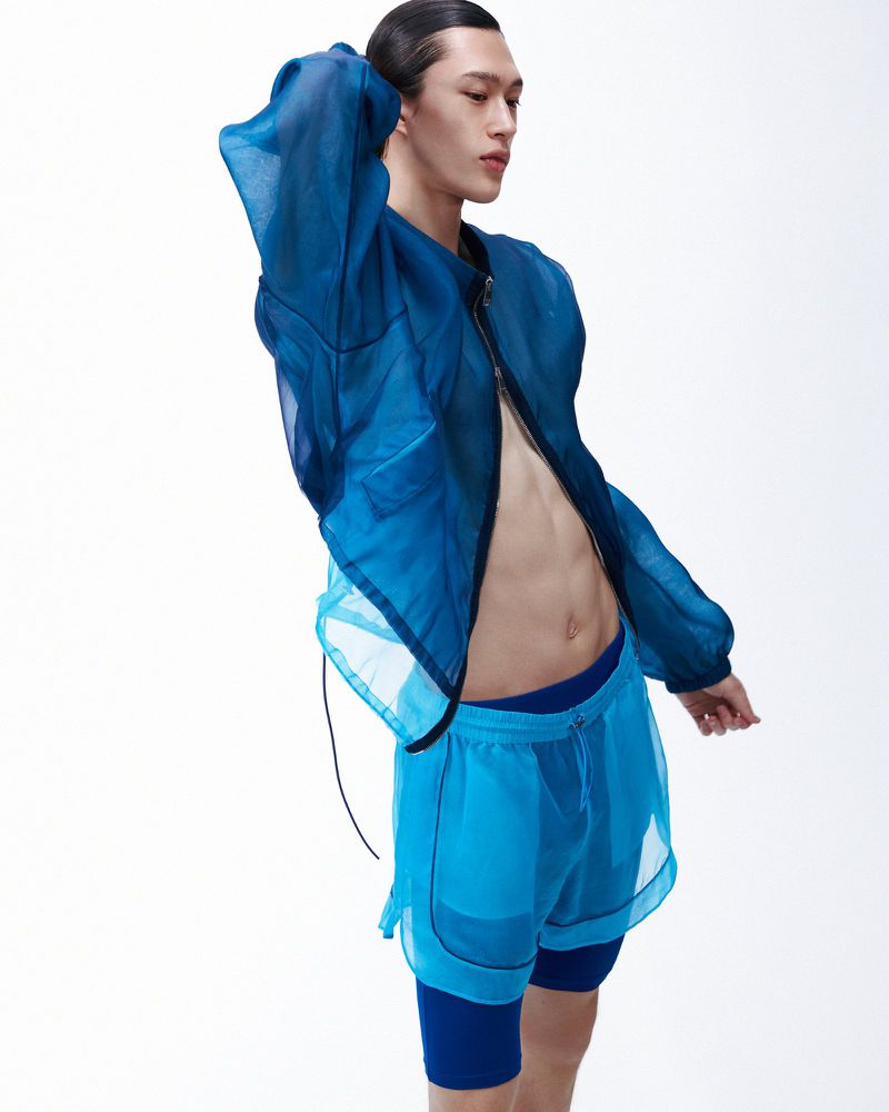 Model wearing a sheer blue zip-up jacket and matching athletic shorts against a plain white studio background.