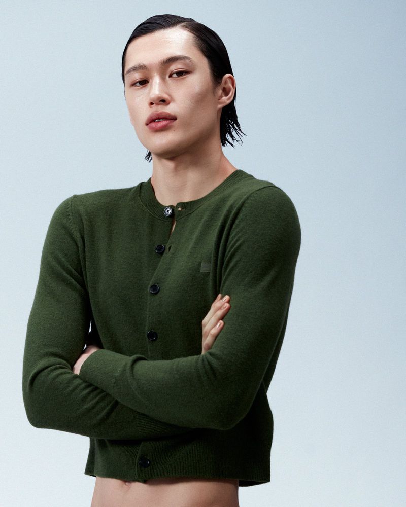 Model with slicked-back hair wearing a dark green buttoned cardigan against a pale gradient background.