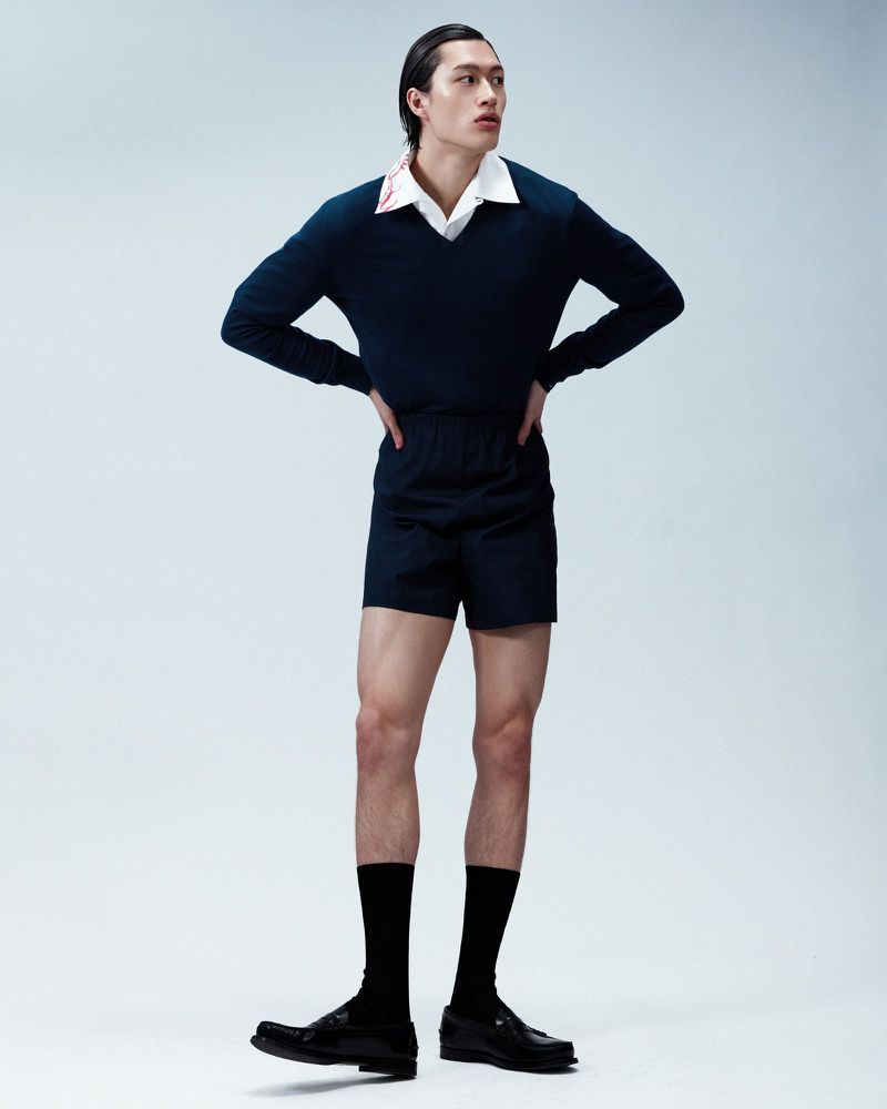 Full-length portrait of a male model wearing a dark navy v-neck sweater, shorts, black socks, and loafers against a light grey background.