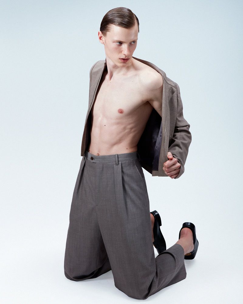 Shirtless male model wearing grey pleated trousers and a taupe jacket kneeling against a white background