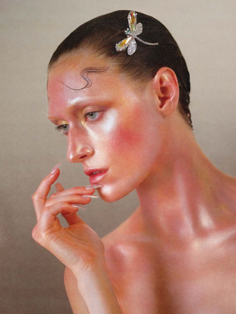 Close-up portrait of a female model with slicked-back dark hair, wearing a jeweled dragonfly hair clip against a neutral grey background.