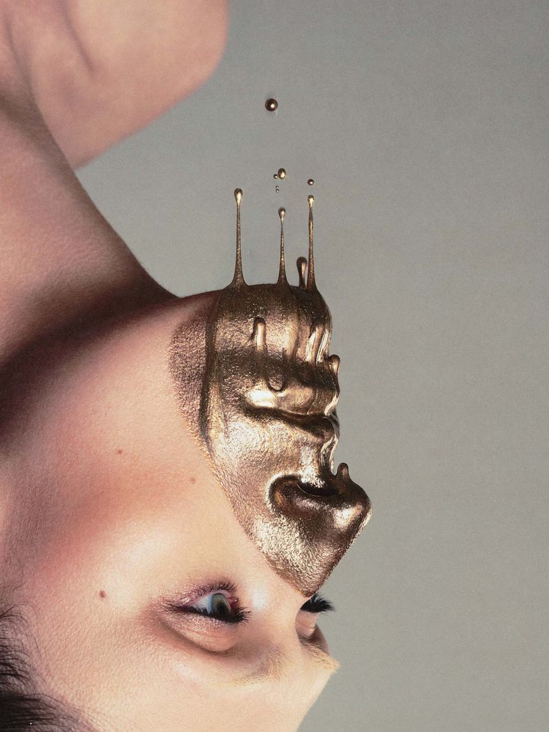 Close-up portrait of a model with metallic gold liquid dripping from the face against a neutral grey background.