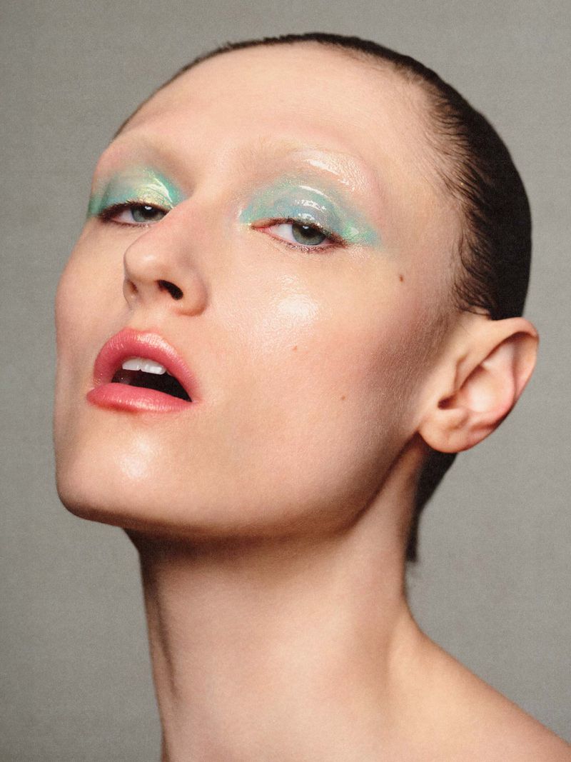 Close-up beauty portrait of a model with glossy green eyeshadow and pink lips against a plain grey backdrop.