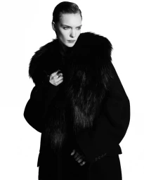 Model Penelope Ternes wearing a dark fur-collared coat against a pure white background in a studio fashion portrait.