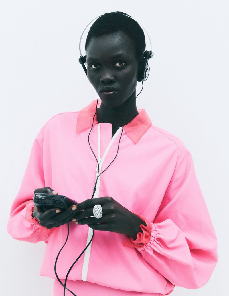 Black woman wearing a pink zip-up jacket and headphones holding a personal music player