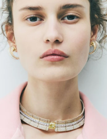 Close-up portrait of a model wearing layered diamond choker necklaces, pavé earrings, and a pink jacket against a neutral studio background.