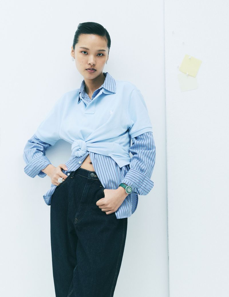Model wearing a layered blue polo shirt and striped button-down over dark jeans against a white seamless background.
