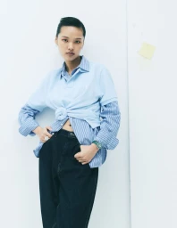 Model wearing a layered blue polo shirt and striped button-down over dark jeans against a white seamless background.