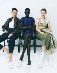 Three female models wearing casual fashion clothing and white slippers seated on black chairs against a white background