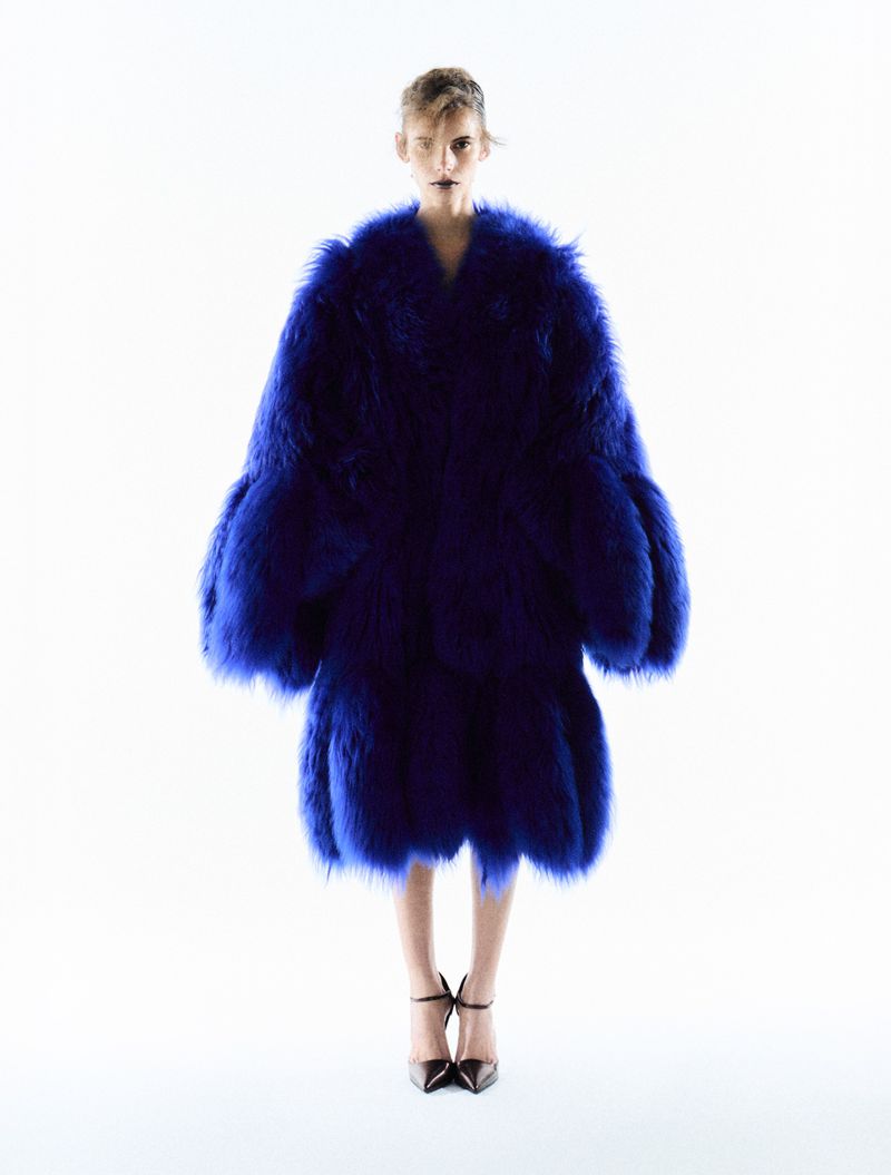 A woman with light hair poses in a floor-length, cobalt blue fur coat and black high heels against a pure white backdrop. Fashion shot.