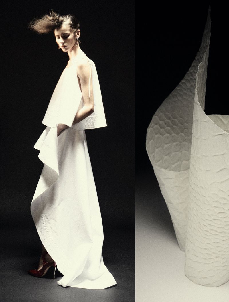 Model in a sculptural white dress with voluminous flared layers standing against a dark background next to paper art sculpture.