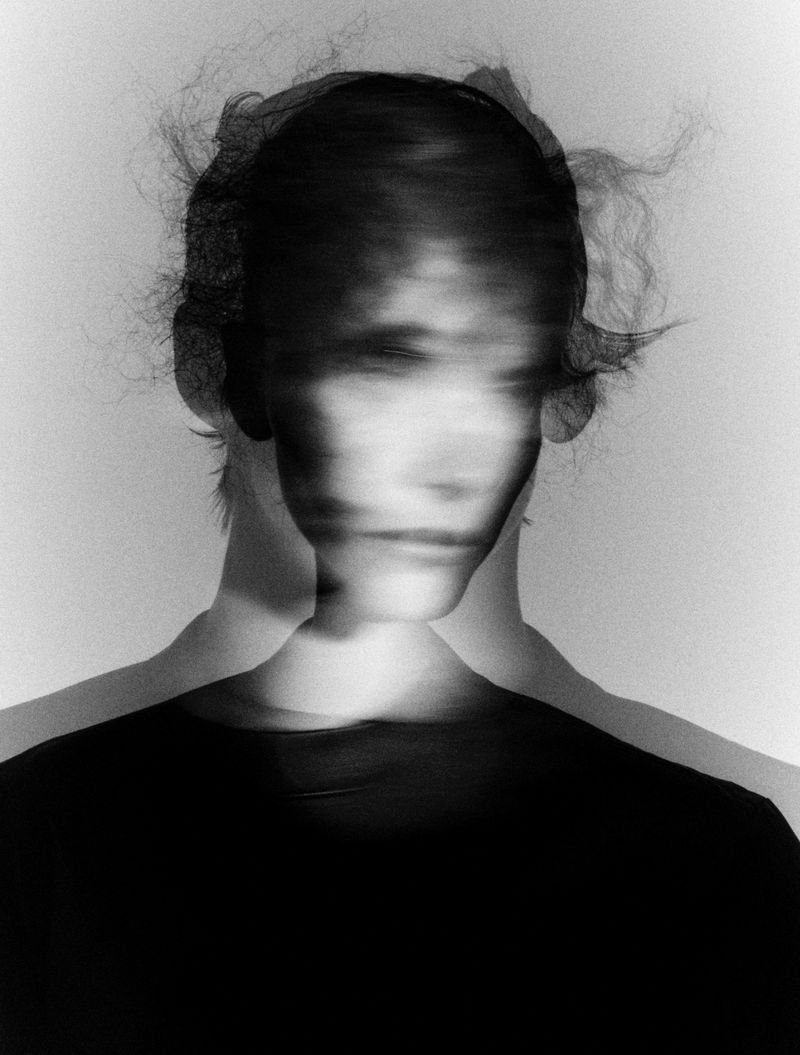 Monochrome portrait of a model with motion blur showing layered overlapping facial features against a light grey background.