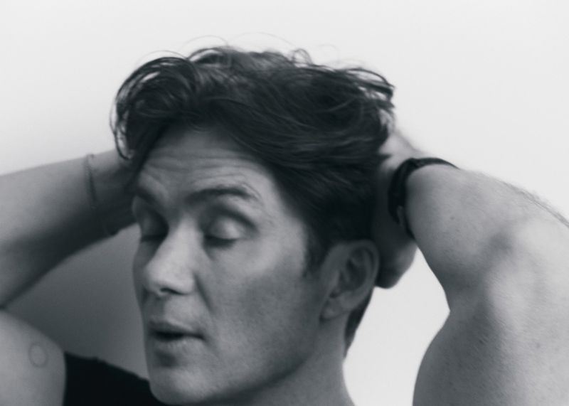 Monochromatic portrait of an adult man with his hands behind his head, against a bright, neutral background in a close-up shot.