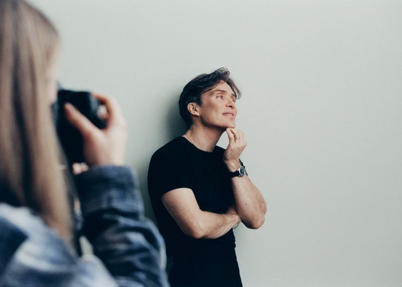 Indoor portrait of a light-skinned adult male in a black t-shirt with hand to chin, photographed by a person with a camera.