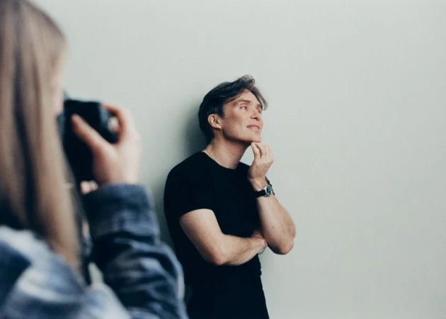 Indoor portrait of a light-skinned adult male in a black t-shirt with hand to chin, photographed by a person with a camera.