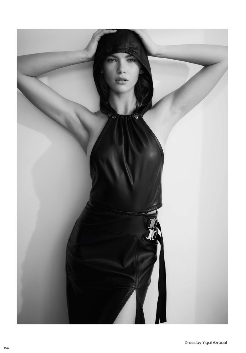 Model wearing a black leather halter top with matching hood and skirt, posing against a white studio wall in monochrome.
