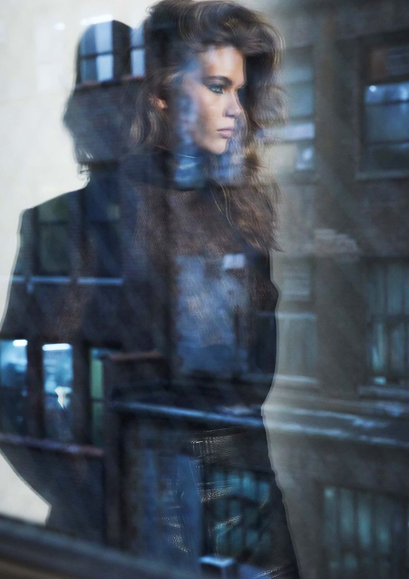 Woman with wavy brown hair and black jacket gazes through window with building reflections, creating a surreal, layered urban portrait.