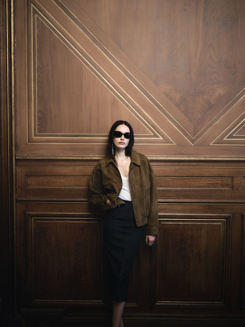 Model wearing dark sunglasses, brown suede jacket, and dark skirt leaning against an ornate wood-paneled wall