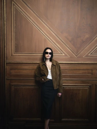 Model wearing dark sunglasses, brown suede jacket, and dark skirt leaning against an ornate wood-paneled wall
