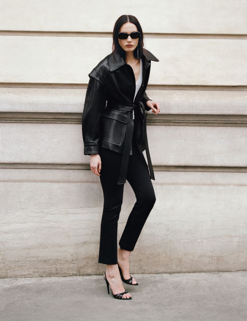 Model wearing a black leather belted jacket and sunglasses standing against a stone wall for Nour Hammour Summer 26.