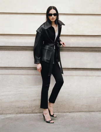 Model wearing a black leather belted jacket and sunglasses standing against a stone wall for Nour Hammour Summer 26.