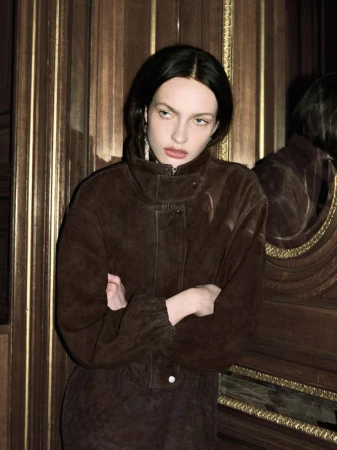 Model in brown suede jacket with arms crossed standing against ornate dark wood paneling