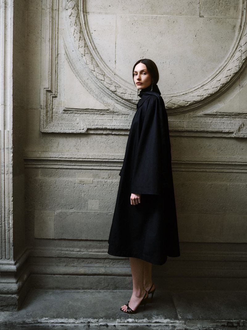Fashionable woman in a black cape stands in front of a textured concrete wall, creating a minimalist and elegant visual statement.